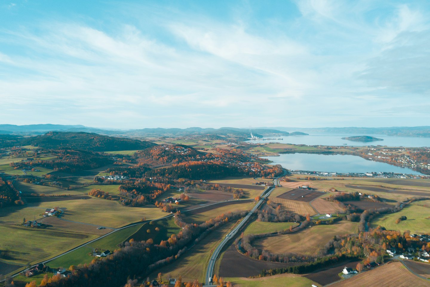 Lufthøydevisning av høstbakker, motorvei og innsjø med fjerne fjell.